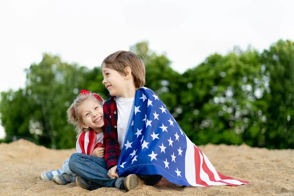 Hermano y hermana en la orilla, envueltos en la bandera estadounidense, niños felices riendo, niña y niño concepto de patriotismo y la celebración de la independencia de Estados Unidos. Día de los Veteranos — Foto de Stock