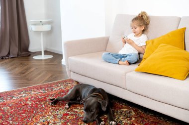 Kanepede oturan küçük sarışın bir kız. Cep telefonu, büyük bir köpek, çocuklar ve aletleri var..