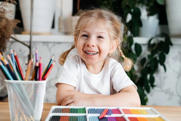 Ένα μικρό ξανθό κορίτσι με ένα γοητευτικό χαμόγελο κάθεται σε ένα τραπέζι με ένα φωτεινό πολύχρωμο παλέτα έγχρωμη πλαστελίνη για το μοντέλο στο σπίτι, χόμπι και την ανάπτυξη των παιδιών — Φωτογραφία Αρχείου