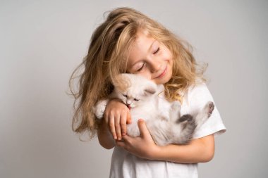 Küçük sarışın bir kız, elini yalayan sevimli beyaz bir kediye sarılıyor, bir çocuk ve bir kedi yavrusu, güzel bir kız gülümsüyor.