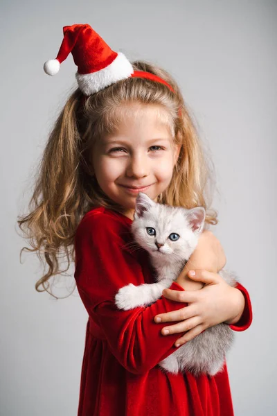 Una niña rubia hermosa en un vestido rojo y un sombrero de Santa con un lindo gatito blanco en sus manos, un niño feliz, el concepto de regalos de Navidad — Foto de Stock