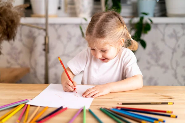 Ένα μικρό ξανθό κορίτσι κάθεται στο τραπέζι, χαμογελώντας και ζωγραφίζοντας μια καρδιά με ένα κόκκινο μολύβι, υπάρχουν πολλά χρωματιστά μολύβια στο τραπέζι, μαθαίνοντας έννοια — Φωτογραφία Αρχείου