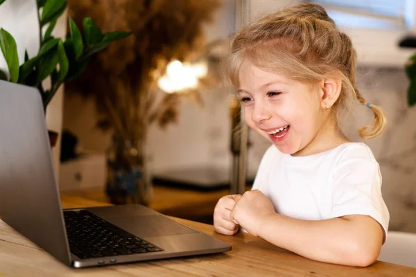 Ένα μικρό ξανθό κορίτσι με ένα φορητό υπολογιστή κάθεται σε ένα τραπέζι και χαμογελά — Φωτογραφία Αρχείου