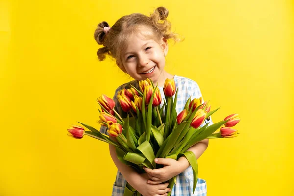 Маленькая блондинка на желтом фоне с букетом красных тюльпанов в руках, смеющаяся. Концепция отдыха — стоковое фото