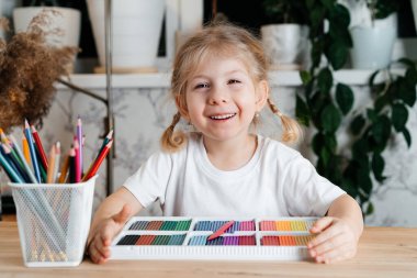 Çekici bir gülümsemesi olan küçük sarışın bir kız, renkli renkli bir palet ile bir masada oturuyor. Evde modellik, hobiler ve çocuk gelişimi için.