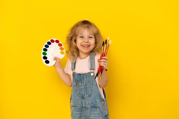 Blondynka kręcona na żółtym tle uśmiecha się z farbami i frędzlami w dłoniach, ubrana w dżinsowy kombinezon i różowy T-shirt — Zdjęcie stockowe