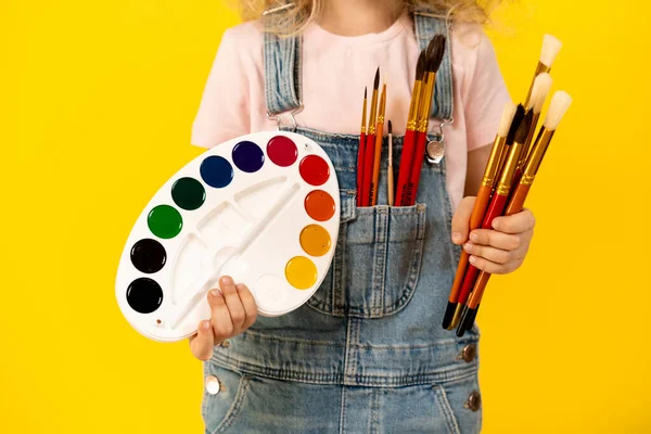 Farbe und Pinsel in den Händen eines kleinen Mädchens auf gelbem Hintergrund, Nahaufnahme, das Konzept der Kreativität und Entwicklung von Kindern — Stockfoto
