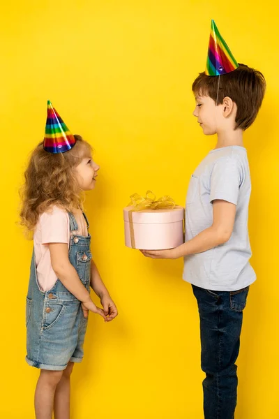 Kleines blondes Mädchen nimmt Geschenk in rosa Schachtel zum Geburtstag von ihrem Bruder an, auf gelbem Hintergrund, Kinder in Mützen, vertikales Foto — Stockfoto