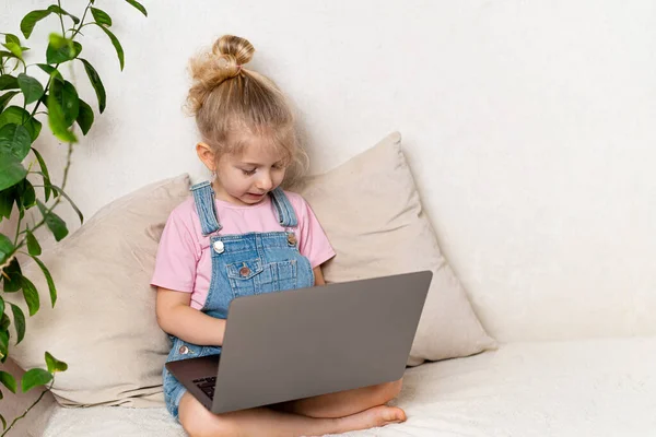 Ευτυχισμένο μικρό ξανθό κορίτσι κάθεται στο σπίτι σε ένα κρεβάτι με ένα φορητό υπολογιστή, ένα μέρος για κείμενο, η έννοια της μάθησης στο σπίτι — Φωτογραφία Αρχείου