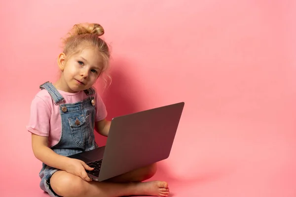 Ένα χαμογελαστό κοριτσάκι κάθεται με ένα φορητό υπολογιστή απομονωμένο σε ροζ φόντο. Εκπαίδευση εξ αποστάσεως στο σπίτι κατά τη διάρκεια της καραντίνας. banner με χώρο για κείμενο — Φωτογραφία Αρχείου