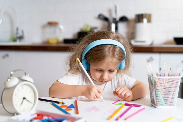 Μικρό ξανθό κορίτσι στο σπίτι στην κουζίνα στο τραπέζι με ακουστικά, ζωγραφίζει και μαθαίνει εξ αποστάσεως — Φωτογραφία Αρχείου