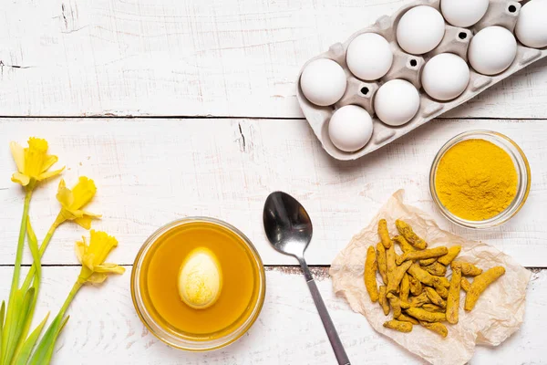 Der Prozess des Bemalens von Ostereiern mit natürlichen Pflanzenfarbstoffen, Kurkuma, auf weißem Holzgrund, Ansicht von Hühnereiern und gelben Narzissen — Stockfoto