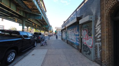 Bronx 'un Unionport bölgesindeki iç mahallenin gerçekliği. Başı örtülü Müslüman bir kadın sokağın karşısında solunda metro treniyle karşıdan karşıya geçiyor ve sağda vandalize dükkanları kapatıyor..