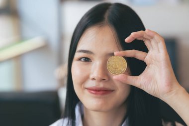 Genç Asyalı kadın gülümsüyor, Bitcoin kriptosu tutuyor. Bitcoin olarak dijital paraya para yatıran Asyalı bir kadın. Genç tüccar ya da yatırımcı Kripto para birimiyle ilgileniyor.