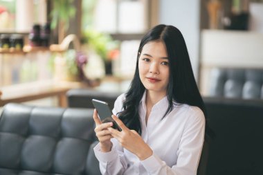 Genç Asyalı bir kadın bir kafede akıllı telefondan mesaj yazıyor. Genç bir kadının kahve masasında cep telefonu kullanırken çekilmiş görüntüsü..