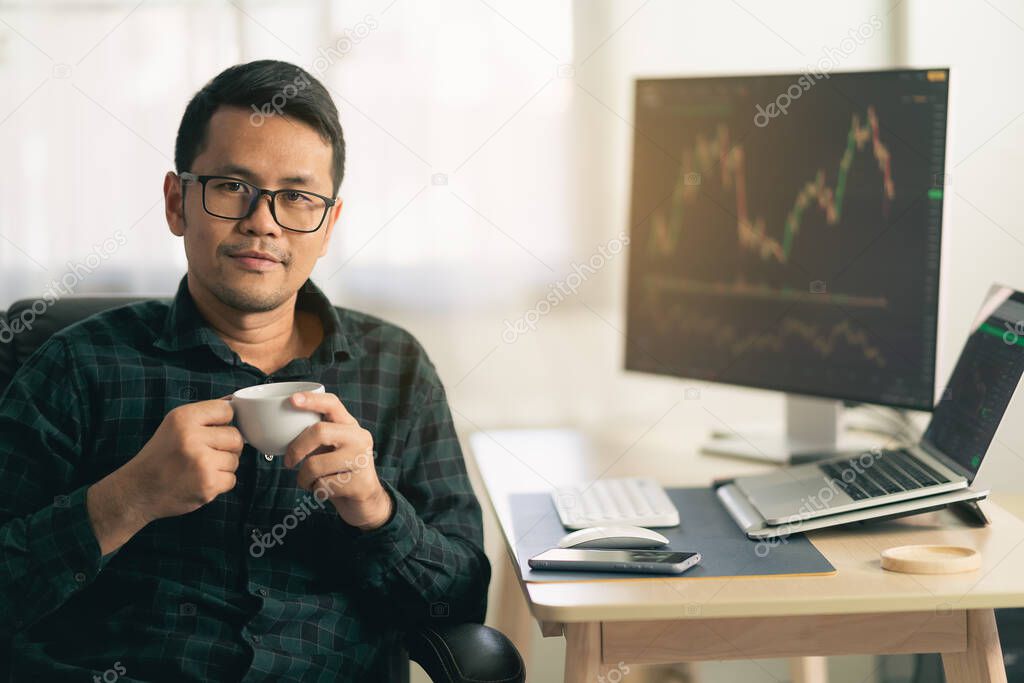 Asian man trader drinking coffee and sitting at home office in front of monitors with cryptocurrency graph holding smartphone browsing application monitoring cryptocurrency price