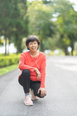 Kıdemli Asyalı kadın koşucu gülümsüyor. Mobil akıllı telefon uygulaması kullanıyor. Fitness motivasyonu için kulaklıkla müzik dinliyor. Asyalı kadın parkta koşuya hazırlanıyor.