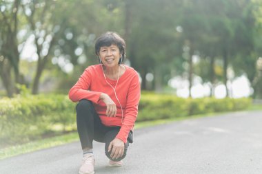 Kıdemli Asyalı kadın koşucu gülümsüyor. Mobil akıllı telefon uygulaması kullanıyor. Fitness motivasyonu için kulaklıkla müzik dinliyor. Asyalı kadın parkta koşuya hazırlanıyor.