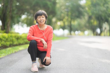 Kıdemli Asyalı kadın koşucu gülümsüyor. Mobil akıllı telefon uygulaması kullanıyor. Fitness motivasyonu için kulaklıkla müzik dinliyor. Asyalı kadın parkta koşuya hazırlanıyor.