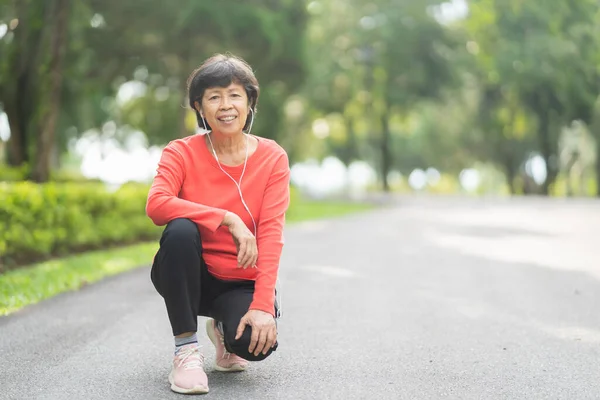 Kıdemli Asyalı kadın koşucu gülümsüyor. Mobil akıllı telefon uygulaması kullanıyor. Fitness motivasyonu için kulaklıkla müzik dinliyor. Asyalı kadın parkta koşuya hazırlanıyor.