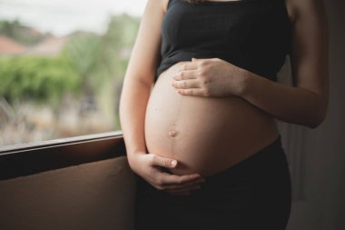 Asyalı hamile kadın ellerini şişmiş karnında tutuyor. Hamile bir kadın evde mutlu bir aşk yaşıyor. Doğum öncesi bakım ve kadın kalp hamileliği..