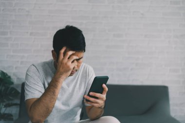 Depresyon kavramı. Asyalı adam üzgün ve cep telefonunu kontrol ediyor. Oturma odasındaki koltukta oturuyor. Asyalı adam ciddi ve cep telefonu kullanırken endişeleniyor..