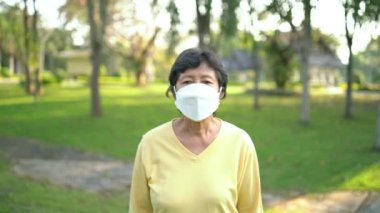 60 'lı yaşlardaki atletik Asyalı kadın cerrahi maske takıyor, gülümsüyor ve koşuyor. Güneşli bir günde parkta koşan güzel, kıdemli Asyalı kadın..