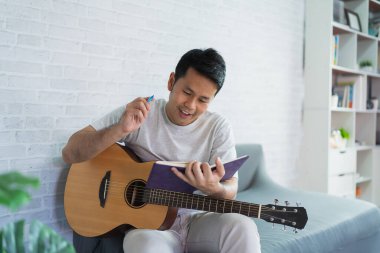 Aydınlık oturma odasında koltukta oturan Asyalı adam gitar çalıyor. Asyalı adam evde gitar çalarken şarkı yazıyor. Şarkı müziği konsepti oluştur.