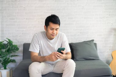 Oturma odasındaki kanepede oturan sıradan giyinmiş gülümseyen Asyalı adam. Asyalı adam oturma odasında cep telefonu sohbeti yapıyor..