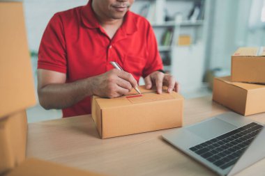 Kırmızı üniformalı Asyalı kurye evde çalışıyor, paket kutularını posta yoluyla gönderme veya taşıma için kontrol ediyor.