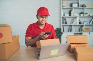 Asian delivery man worker smiling using mobile scan barcode for boxes for sending or conveying parcels by mail. Wearing red uniform work at home.