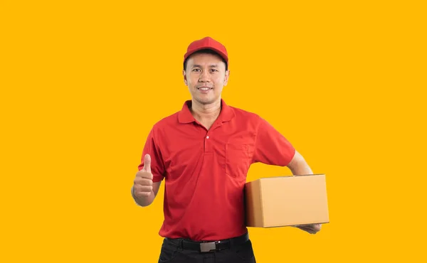 亚洲送货员笑着竖起大拇指 身穿红色制服 背景黄色 手持包裹盒 用于邮寄或运送包裹 — 图库照片