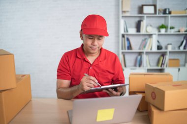 Kırmızı üniformalı Asyalı teslimatçı evde çalışıyor, paket kutularını posta yoluyla gönderme veya taşıma için kontrol ediyor.