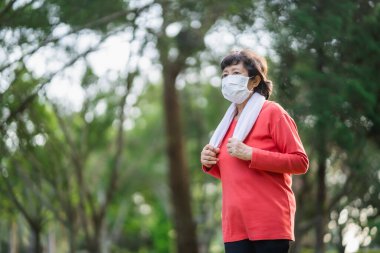 60 'lı yaşlarda, cerrahi maske takıp koşu yapan Atletik Asyalı bir kadın. Güneşli bir günde parkta koşan güzel, kıdemli Asyalı kadın..