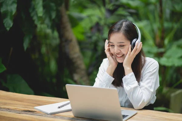 幸せな若いアジアの女性はノートパソコンを使って笑顔で白いヘッドフォンをしてカフェに座っています 若いアジア人女性座っていますでコーヒーショップとビデオ通話会議上のラップトップ — ストック写真