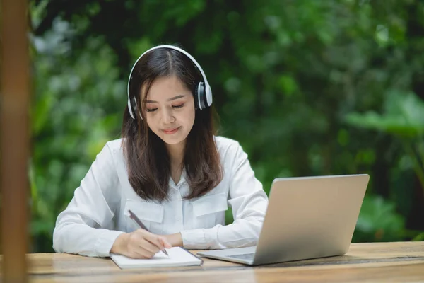 ノートパソコンを使って白いヘッドフォンをつけてノートを書く幸せな若いアジアの女性の笑顔 若いアジア人女性座っていますでコーヒーショップとビデオ通話会議上のラップトップ — ストック写真