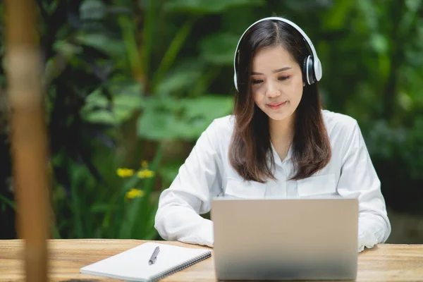 幸せな若いアジアの女性はノートパソコンを使って笑顔で白いヘッドフォンをしてカフェに座っています 若いアジア人女性座っていますでコーヒーショップとビデオ通話会議上のラップトップ — ストック写真