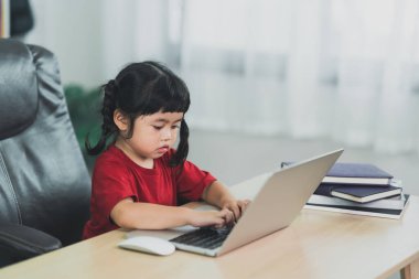 Asian baby girl wearing a red t-shirt use laptop and study online on wood table desk in livingroom at home. Education learning online from home concept.