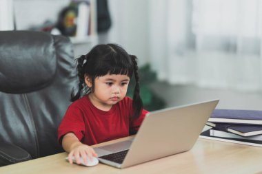 Asian baby girl wearing a red t-shirt use laptop and study online on wood table desk in livingroom at home. Education learning online from home concept.