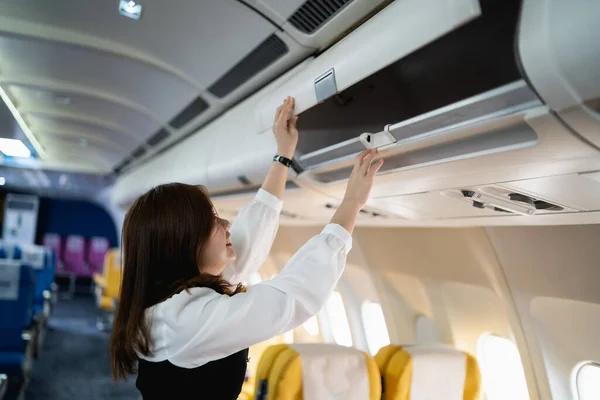 Asian young woman open the top shelf to get luggage on airplane.Cabin crew girl lift luggage bag in airplane . Airline transportation and tourism concept.