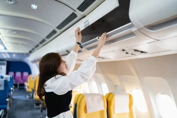 Asian young woman open the top shelf to get luggage on airplane.Cabin crew girl lift luggage bag in airplane . Airline transportation and tourism concept.