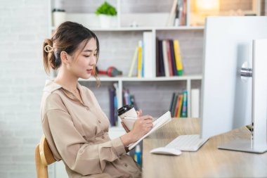 Asian freelance woman smiling holding cup of hot coffee and working on laptop computer on wooden table at home. Entrepreneur woman working for her business at home. Business work at home concept.