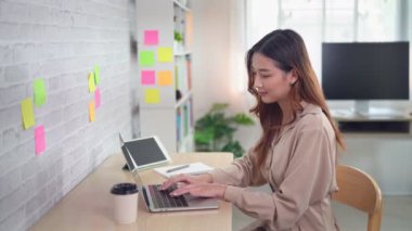 Asian freelance woman smile typing on keyboard and working on laptop on wooden table at home. Entrepreneur woman working for her business at living room home. Business work at home concept.