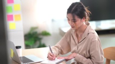 Asian freelance woman smiling using pen making notices in her notebook and working on laptop on wooden table at home. Entrepreneur woman working for her business at home. Business work at home concept