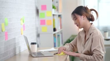 Asian freelance woman smiling paste post-it on the wall to record for work and using on laptop on wooden table at home. Entrepreneur woman working for her business at home. Business concept.