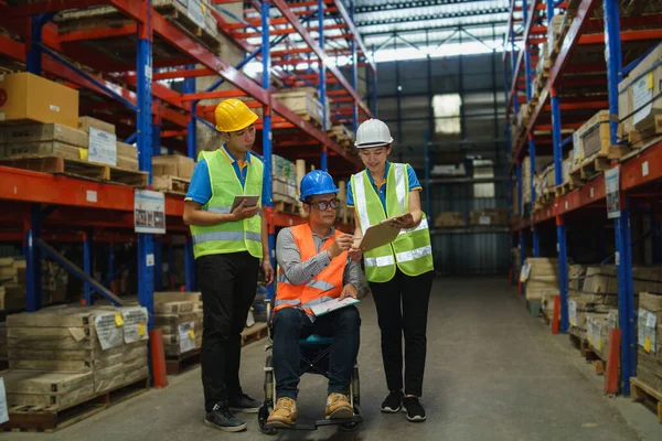 Manager handicapped in wheelchair man employee using tablet check stock work at warehouse. Worker wearing high visibility clothing and hard hat, helmet and checking count up goods boxes for delivery.