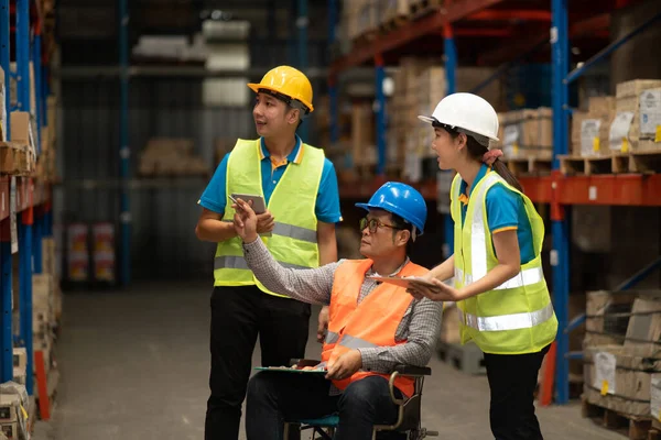 Manager handicapped in wheelchair man employee using tablet check stock work at warehouse. Worker wearing high visibility clothing and hard hat, helmet and checking count up goods boxes for delivery.