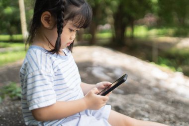 Asian baby girl smiling and using mobile selfie make a photo picture in the park garden. Cute girl learning to use smartphone by her self. Education learning baby concept.