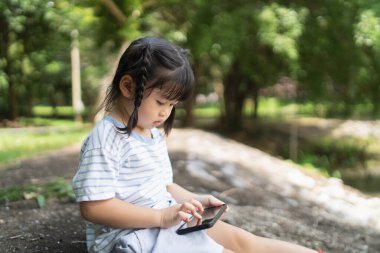 Asian baby girl smiling and using mobile selfie make a photo picture in the park garden. Cute girl learning to use smartphone by her self. Education learning baby concept.