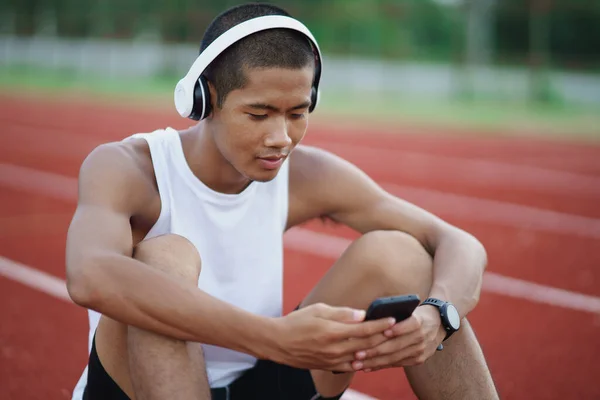 Asian man sportsman fit body wearing white headphone and using mobile phone to listen music while resting or before jogging, exercising at lane stadium. Sport healthy running concept.